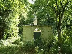 Photographie en couleurs d'une maisonnette ruinée envahie par la végétation d'un sous-bois.