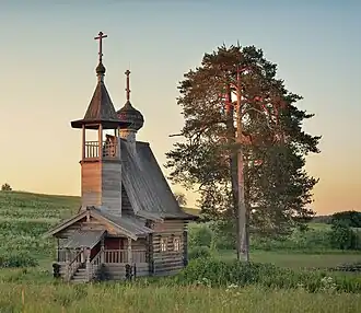 Vue au couchant d'une chapelle en bois qui possède un petit beffroi et un toit en tente, le tout d'une architecture en bois russe.
