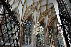 vue en contre plongée et à travers une grille entrouverte de l’intérieur d’une chapelle, avec quatre grandes fenêtres dotées de vitraux et une voûte réticulée.