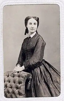 Charlotte photographiée en noir et blanc a l'air mélancolique, elle se tient debout, les mains posées sur le dossier d'un fauteuil capitonné