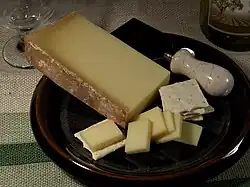 La photographie en couleur représente une tranche de beaufort dans une assiette brune, posée sur une nappe à gros grain. Dans l'assiette se trouvent aussi des tranchettes de fromage, des crackers et un couteau à fromage large en inox à manche en marbre ; un exemple de service du beaufort à l'apéritif