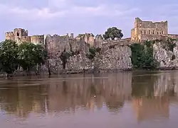 .Château de vielles pierres en ruine, formé de murs bruts et de larges tours, s'étalant en longueur au sommet d'une petite falaise, au pied de laquelle coule une rivière. Le ciel est bleu pastel et le château est rosé, dû à l'heure matinale à laquelle la photo a été prise.