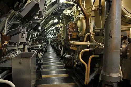 Un long couloir avec sur la droite de nombreux tuyaux et sur la droite une cloison qui s'incurve vers l'intérieur