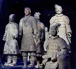 Photographie de six statues de soldats en terre cuite coiffés et vêtus distinctement, l'un assis au premier plan et les autres debout.