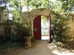 Photographie en couleurs d'un espace végétalisé limité par des murs et un portail en bois.