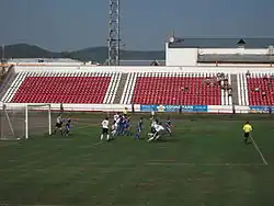 Photographie de la pelouse d'un stade de food centré au niveau d'un but, avec des joueurs jouant au foot devant. En arrière-plan se trouve des petits gradins colorés en rouge et blanc.