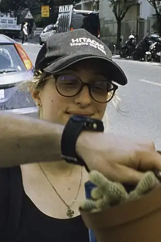 une jeune femme avec des lunettes et une casquette, dans une rue. Une main passe en partie devant son visage