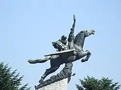 statue de cheval ailé vue de profil, placée en hauteur. L'animal est tendu en avant avec deux personnes sur le dos, la première personne lève un bras en l'air