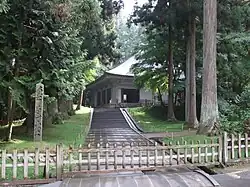Photo couleur d'un chemin de dalles grises terminé par un escalier de pierres grises au pied d'un bâtiment à un étage d'un temple bouddhique et bordé d'étendues de pelouses boisées. Au premier plan, une clôture en bois ferme l'accès du chemin, et, à l'arrière-plan, on distingue une forêt sous un viel blanc laiteux.
