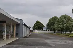 Autour d'une cour goudronnée, deux bâtiments techniques (aux toits d'usine) se font face. À droite, des bosquets d'arbre. À gauche, au premier plan, on distingue le porche d'une cour couverte.