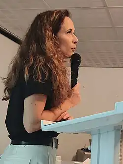 photo de profil d'une femme parlant depuis un pupitre, micro en main.