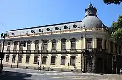 Photographie sous un ciel bleu azur d'un bâtiment de couleur crème à deux étages et à toit mansardé et à neuf travées.