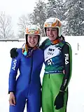 Elena Runggaldier en combinaison de saut à ski bleue, se tenant par les épaules avec Evelyn Insam en combinaison verte et dossard de compétition, toutes deux casquées et souriantes, sur fond de paysage hivernal avec des sapins enneigés