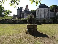 Photographie en couleurs d'un terrain enherbé avec un ancien puits au centre et un bâtiment en arrière-plan.