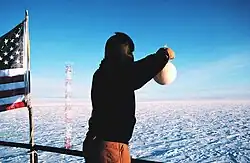 Photo en couleur. En extérieur, un homme de dos, le visage emmitouflé dans une cagoule, tend le bras au-dessus d'une rambarde, une fiole à la main. À sa gauche, un drapeau américain. En arrière plan et jusqu'à l'horizon, une étendue de glace ou de neige.