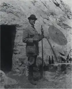 Photographie en noir et blanc d'un homme se tenant devant l'entrée d'une grotte, un bâton ferré à la main.