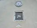 Au milieu est dessiner une colombe avec au bec un rameau d'olivier (symbole de paix), au-dessus une horloge, et au-dessous encastrer dans le mur, une pierre en marbre en forme de triangle où est sculpté un visage de femme avec un collier