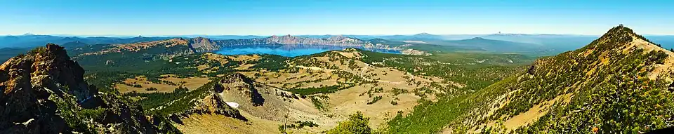 Panorama depuis un sommet vers la caldeira et son lac, dans un paysage d'éboulis, de pelouses alpines et de forêts.