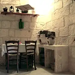 La photographie couleur montre une cuisine en pierre blanchie à la chaux. Une table couverte d'un nappe blanche et deux chaises occupent la moitié gauche. La droite est meublée d'un plan de travail portant des braseros de cuisson. En dessous, des paniers sont rangés. Au-dessus de la table, une étagère porte des lampes anciennes, un moulin à café et un vase.