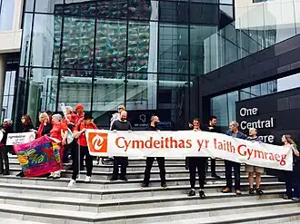 Photo d'une quinzaine de personnes se tenant sur des marches à l'entrée d'un immeuble de bureaux. Ils brandissent des pancartes et tiennent une grande banderole portant l'inscription « Cymdeithas yr Iaith Gymraeg ».