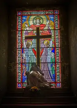 Une Pietà. En arrière-plan, le vitrail représente le Christ Roi avec devant lui, la croix tenue par deux anges.