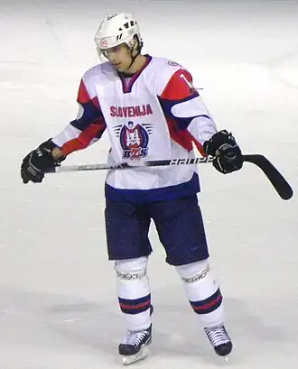 Photographie couleur d’un hockeyeur, de buste, légèrement de profil, le regard dans le vague, portant un maillot blanc avec les épaules vertes claires
