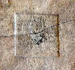 Detail plat d'une pierre beige de gris sur lequel est gravé un carré avec des caractères cunéiformes. La stèle est traversée d'une fente.