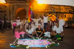 une quinzaine de personnes de couleur mate de peau souriant avec un drapeau transgenre à leurs pieds
