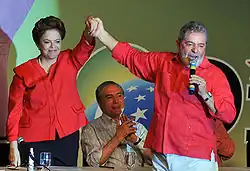 Dilma e Lula vestidos de vermelho. Lula está com um microfone em uma mão e erguendo o braço da Dilma com a outra.