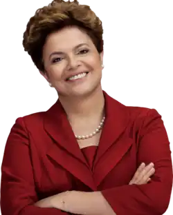 Une femme souriante aux cheveux courts, châtains clairs croise les bras. Elle porte un chemisier bordeaux et un collier de perles.