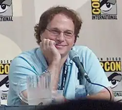 Photographie du buste d'un homme accoudé à un bureau face à un microphone. Il porte des lunettes et une chemise bleue.