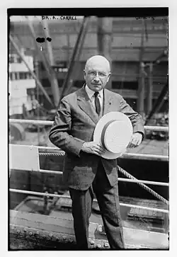 Photo en pied d'un homme en costume tenant un chapeau, posant devant un chantier.