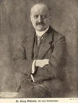Photo en noir et blanc d'un homme âgé assis en costume les mains croisées, il porte une moustache et a le crane dégarni