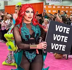 Photographie d'une drag queen blanche souriante sur un tapis rouge. Elle tient une pancarte où est écrit, en gros caractères, "vote vote vote"