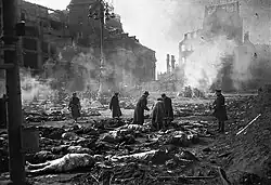Photo noir et blanc montrant une rue en ruine et plusieurs personnes debout parmi des cadavres.