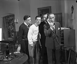Photo en noir et blanc d'une scène de théâtre. Un homme, en costume avec noeud papillon, est au téléphone, trois autres hommes se penchent vers lui pour écouter.