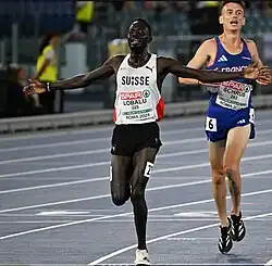 Dominic Lobalu, maillot blanc de la Suisse, écarte les bras en franchissant la ligne d'arrivée.