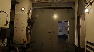 Porte blindée fermant l'entrée des munitions ; lumière du jour en fond, porte du poste de garde sur la droite.