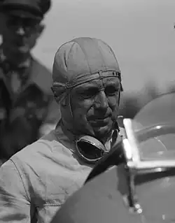 Photo en plan américain de Francis Curzon, en tenue de pilote et au volant d'une voiture.
