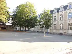 Photographie présentant une cour d'école arborée. Au fond à gauche, un préau. À droite un bâtiment de deux étages (dont un mansardé) sur rez-de-chaussée surélevé, avec un perron de 4 marches devant les portes vitrées.