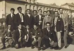 Photo en noir et blancs d'une dizaine de jeunes hommes qui posent en groupe. Ils portent des costumes, et on distingue un bâtiment de leur école à l'arrière-plan
