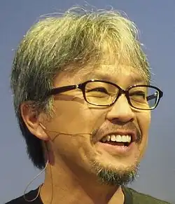 Homme d'origine japonaise portant des lunettes. Ses cheveux et sa barbe sont de couleur grise.