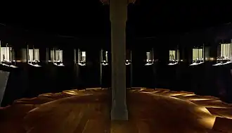 Photographie en couleurs d'une pièce circulaire à l'éclairage tamisé et indirect, des artefacts nichés au sein de vitrines dressées sur le pourtour, un pilier en bois s'élevant au centre de la salle.