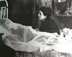 Photographie en noir et blanc d'une femme morte allongée sur un lit, de profil, couverte d'un voile blanc.