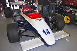 Photographie d'une monoplace de Formule 1 blanche, rouge et bleue, vue de trois-quarts, dans un stand d'exposition.