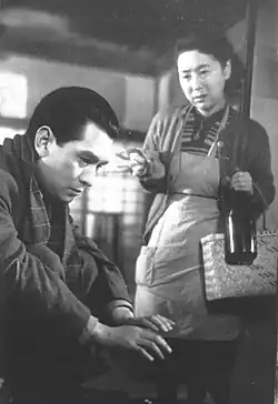 Au premier plan un homme (Ken Uehara), une écharpe autour du cou et l'air sombre, est accroupi et se réchauffe les mains à un brasero, en arrière plan, une femme (Kinuyo Tanaka) pauvrement vétue d'un tablier et d'un gilet, porte un sac en osier et une grande bouteille en verre (image en noir et blanc).