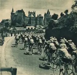 Photo en noir et blanc d'un peloton cycliste se dirigeant vers l'entrée d'une ville avec un château en arrière-plan.