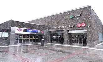 Photo couleur de l'entrée aux portes vitrées d'une station de métro, un bâtiment d'un étage, en briques marrons et oranges, sous un ciel gris. Sur le haut de façade de l'entrée, les inscriptions « metro » et « 9 Paco de Lucía » sont visibles.