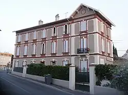 Maison ancienne avec briques rouges en façade, sur trois niveaux, mur de clôture, portail et haie.