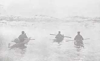 Photographie en noir et blanc de trois hommes chacun embarqué sur un kayak et ramant à proximité d'un littoral.